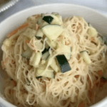 A white bowl filled with thin rice noodles, julienned carrots, and diced cucumbers sits on a table with plates in the background.