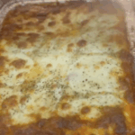 A golden-brown baked lasagna in a foil tray, topped with melted cheese and sprinkled herbs, sits in an oven.