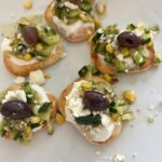 Five crackers topped with cream cheese, chopped cucumber, corn, feta cheese, and a Kalamata olive, arranged on a white plate.
