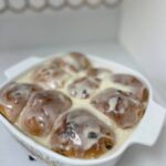 A white dish holds eight iced cinnamon rolls with raisins, freshly baked and covered in glaze, sitting on a white countertop with a honeycomb-patterned backsplash in the background.