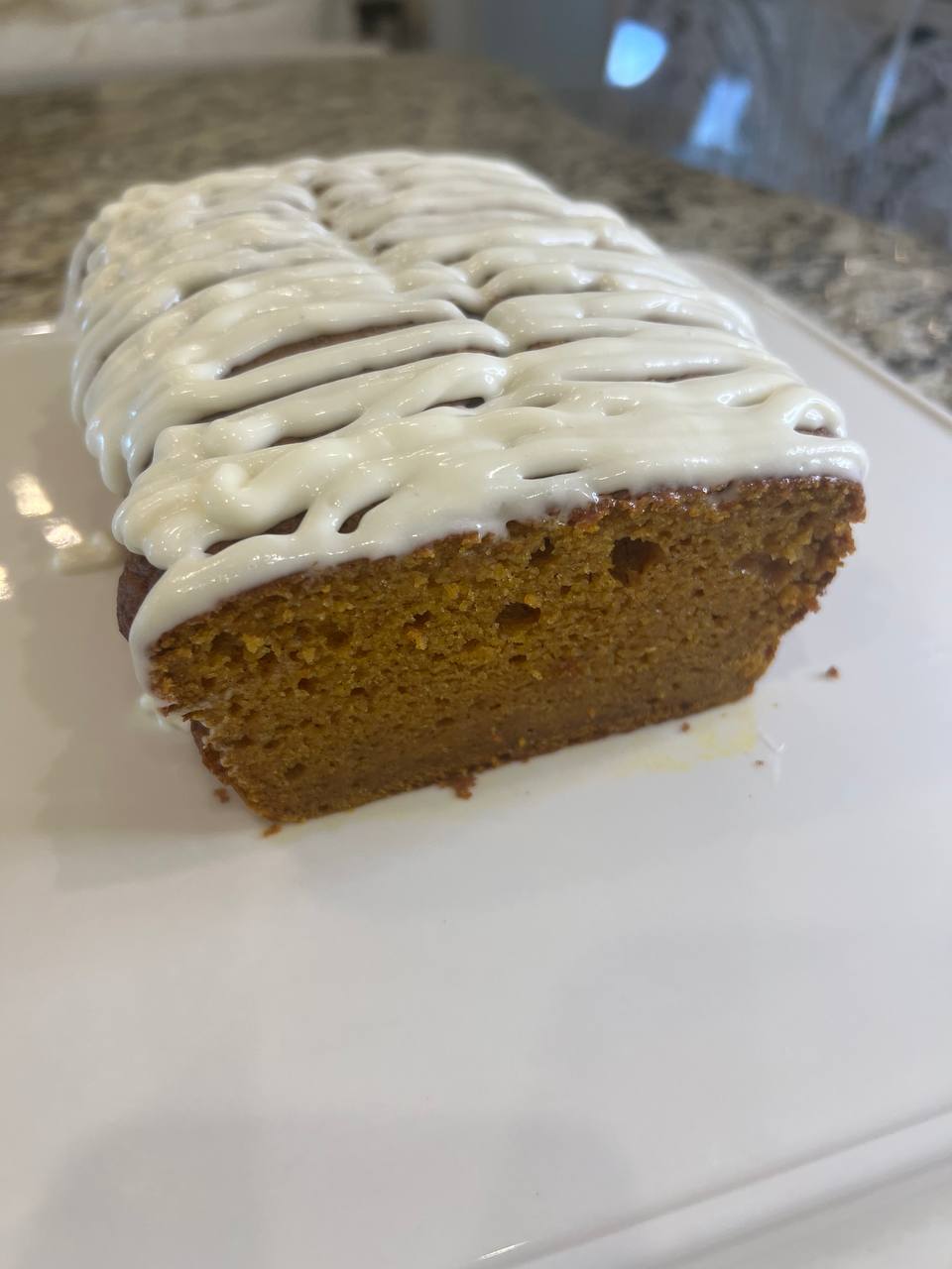 A loaf of pumpkin bread with white icing drizzled on top, sitting on a white plate on a granite countertop.