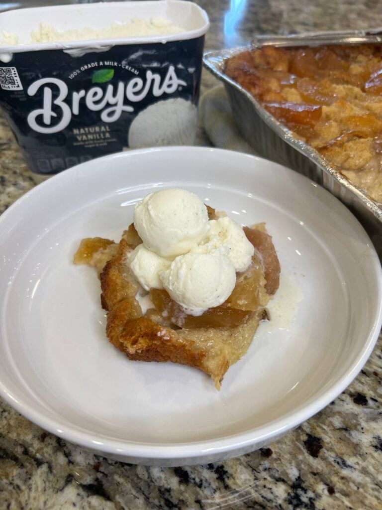 A slice of peach cobbler topped with vanilla ice cream sits in a white bowl. Behind it are a container of Breyers vanilla ice cream and a tray of baked peach cobbler on a granite countertop.