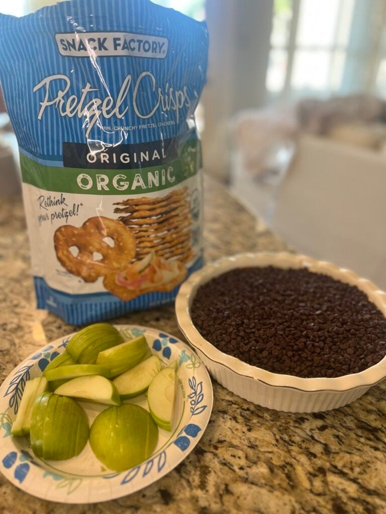 A bag of Snack Factory Organic Pretzel Crisps, a plate of sliced green apples, and a baking dish filled with chocolate chips on a granite countertop.